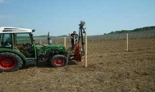 Eine Person bedient einen grünen Traktor mit roten Rädern und setzt mit einer Maschine Holzpfähle auf einem großen, gepflügten Feld unter freiem Himmel ein. Im Hintergrund sind weitere Pfosten und offenes Land zu sehen.