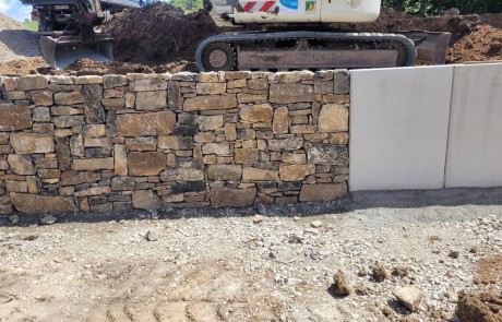 Eine Steinstützmauer geht in einen Betonabschnitt über, während im Hintergrund Baufahrzeuge und Erdbewegungsmaschinen auf einem Erdhügel arbeiten. Im Vordergrund bedecken Schotter und Erde den Boden.