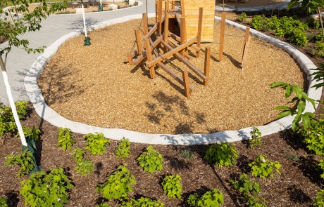 Ein kleiner Spielplatz mit hölzernen Klettergerüsten befindet sich in einem mit Holzspänen bedeckten Kreis, umgeben von einem Gartenbeet mit Grünpflanzen und einem gepflasterten Weg.