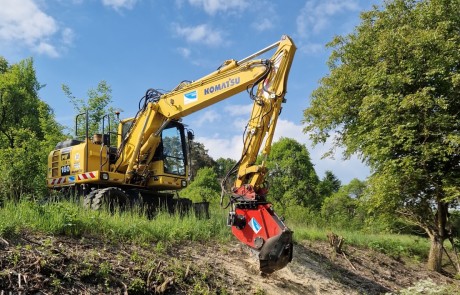 Ein gelber Komatsu-Bagger mit rotem Anbaugerät arbeitet auf einer grasbewachsenen, abschüssigen Fläche in der Nähe von Bäumen unter einem blauen Himmel mit vereinzelten Wolken.