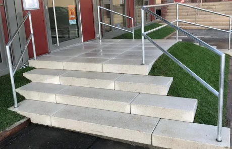 Eine Treppe mit Metallgeländern führt zu einem Gebäudeeingang. Neben der Treppe befindet sich eine Rampe, die jedoch an einer Stufe endet, so dass sie für Rollstühle unzugänglich ist. Gras umgibt die Rampe und die Treppe.